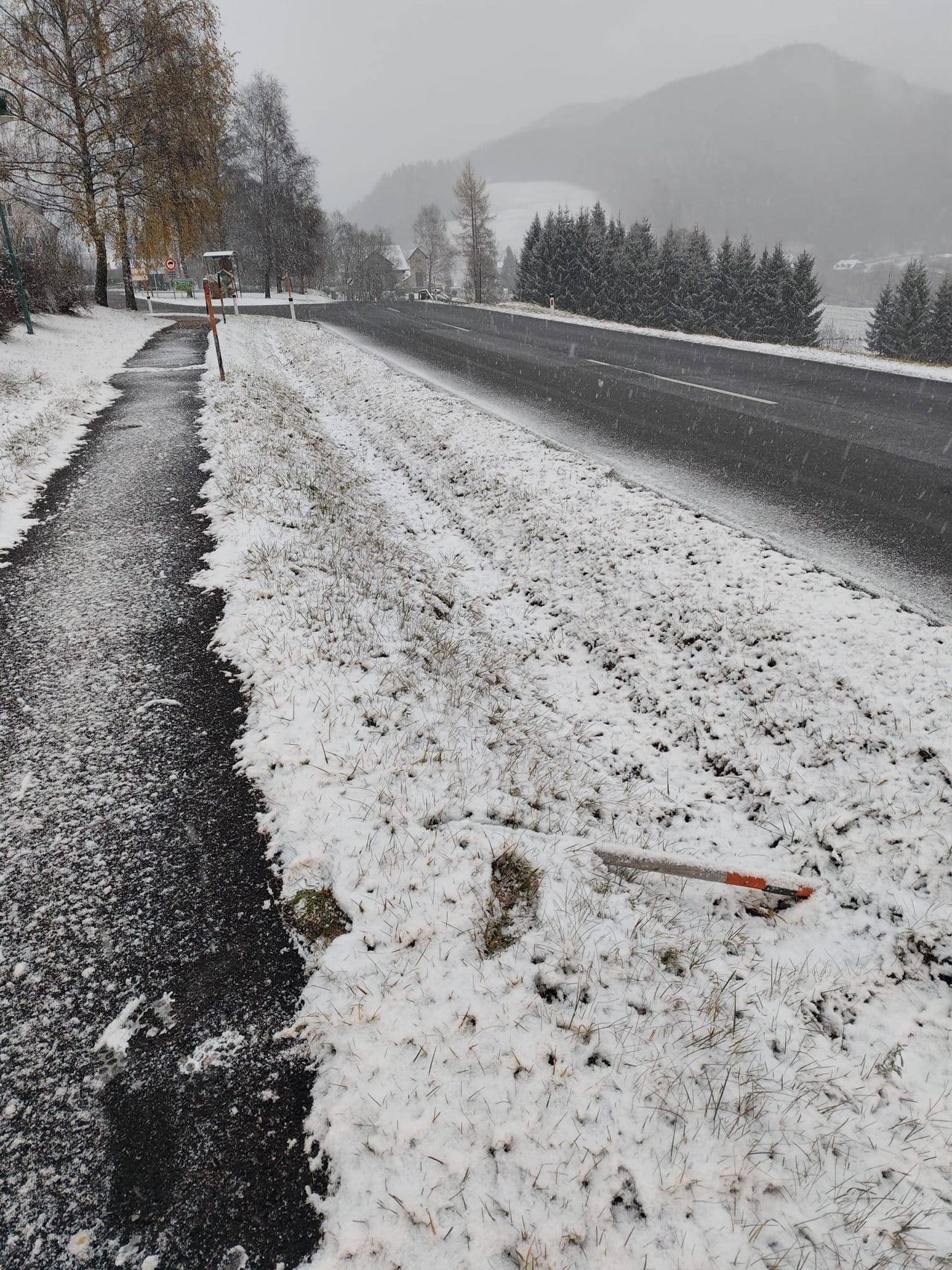 Eine verschneite Straße mit einem Fußweg und Bäumen. Ein Auto fährt auf der Straße, und ein zerbrochener Pfosten liegt an der Seite.