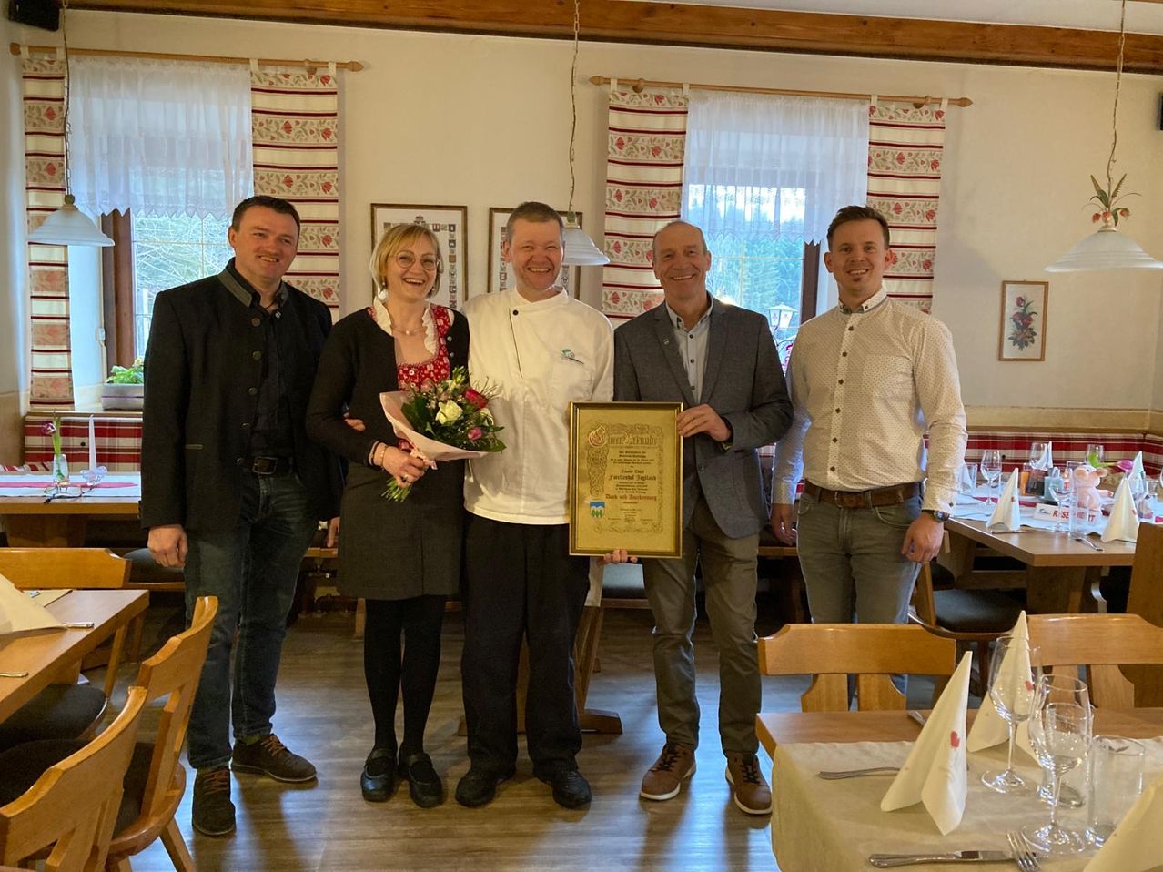 Fünf Personen stehen in einem Raum und lächeln, einer hält eine Urkunde. Eine Frau in der Mitte hält Blumen. Dahinter ist ein Essbereich mit Stühlen und einem Tisch zu sehen.