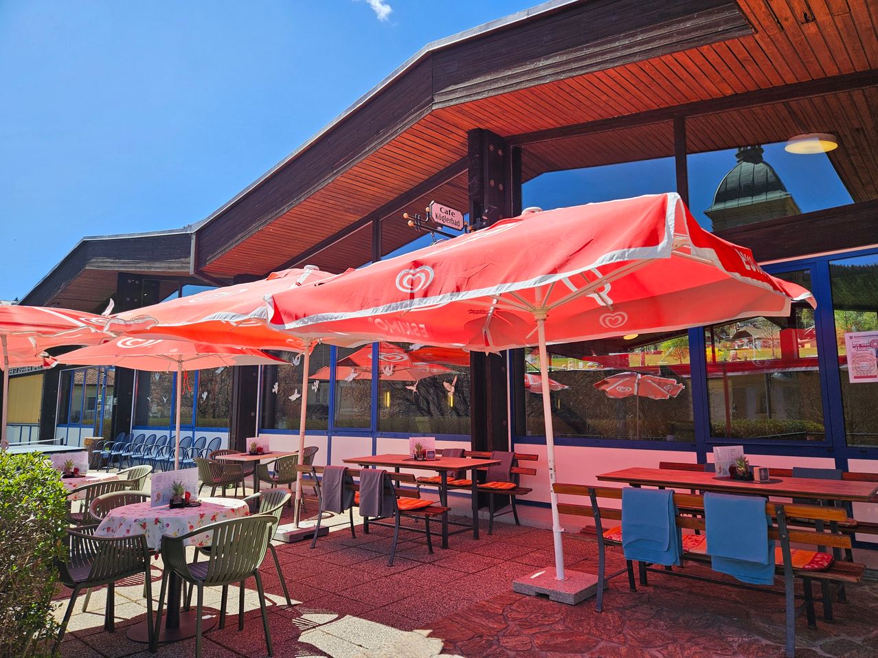 Outdoor-Restaurant mit roten Sonnenschirmen und Tischen. Stühle sind um die Tische mit Tischdecken herum angeordnet. Das Restaurant hat eine Holzüberdachung und Glasfenster.