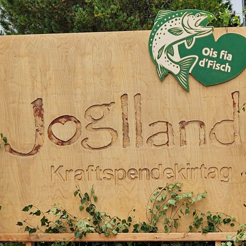 Ein Holzschild mit dem Text Josland Kraftspendekirtag. Es hat ein Fischlogo mit dem Text Ois fia d'Fisch. Am unteren Teil des Schildes wachsen einige Pflanzen.