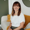 Eine Frau mit braunen Haaren sitzt auf einem Sofa, lächelt und trägt ein weißes Shirt und einen grünen Rock. Sie sitzt auf einem Sofa mit einem gelben Kissen und einem weißen Kissen mit weißen Blumen. Hinter ihr ist eine weiße Wand mit einer grünen Akzentwand und einem Spiegel.