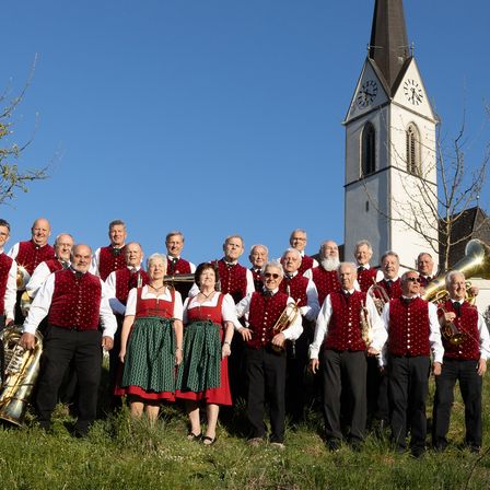Eine Gruppe von Musikern in traditioneller Kleidung posiert vor einer Kirche unter einem klaren blauen Himmel. Sie halten Blechblasinstrumente, darunter Tuba und Trompete.