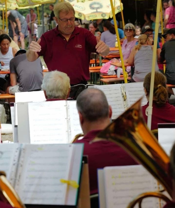A man in a maroon shirt is conducting an outdoor band performance with an audience in the background.