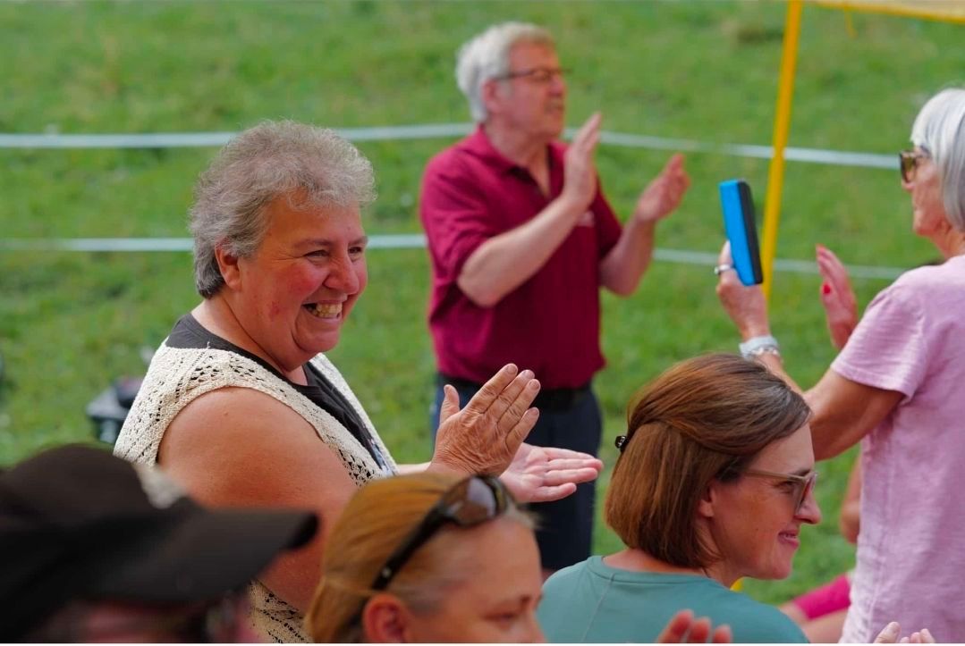 A group of people clapping and smiling in an outdoor setting, with a man in a red shirt and a woman in a white blouse in the foreground.