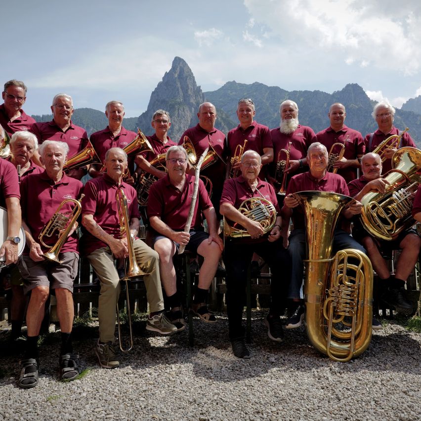 A group of musicians in red shirts pose for a photo with their brass instruments in front of a mountainous backdrop.