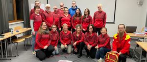 Eine Gruppe von Personen in roten Jacken posiert für ein Foto in einem Raum. Einige sitzen, andere stehen. Medizinische Ausrüstung liegt auf dem Boden.