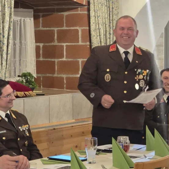 Ein Mann in Uniform steht und lächelt, hält ein Papier in der Hand, umgeben von zwei anderen Männern in Uniform an einem Tisch.