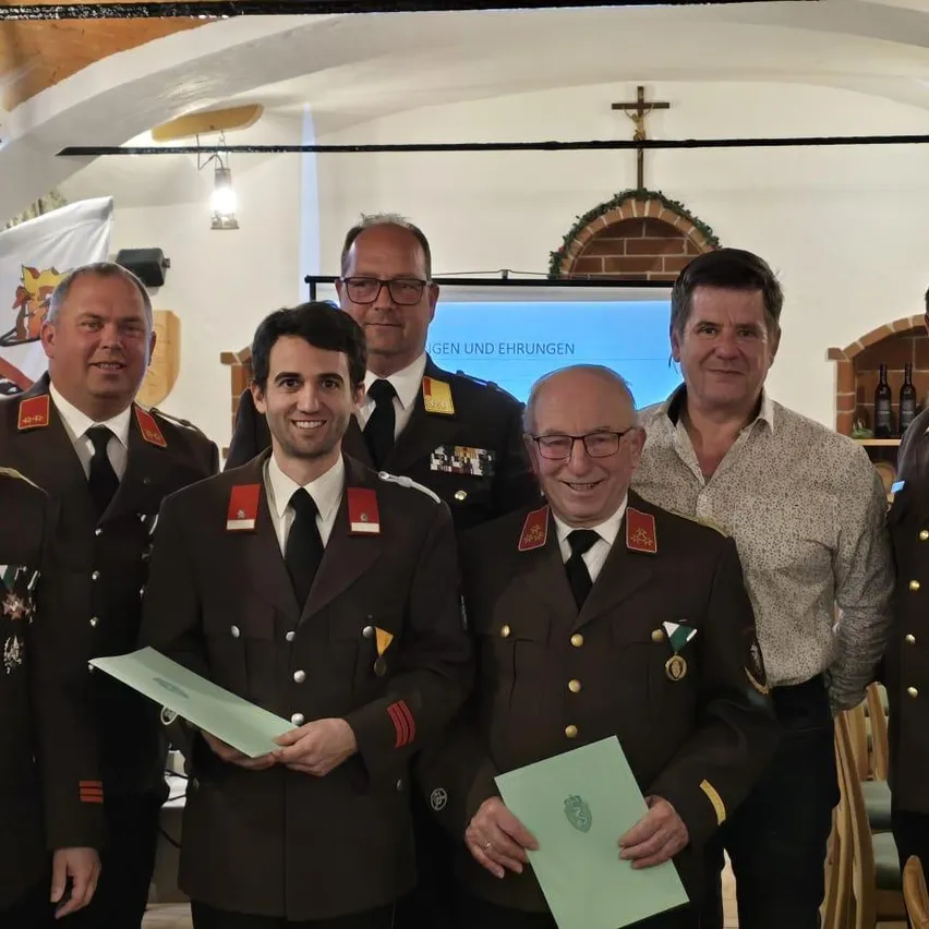 Eine Gruppe von Männern in Uniformen und Medaillen posiert für ein Foto mit einem Mann, der ein Zertifikat hält. Ein Mann mit Brille lächelt. Hinter ihnen ist eine Projektionsleinwand.