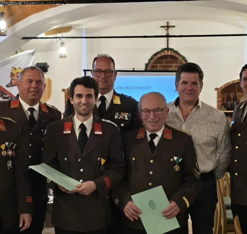 Eine Gruppe von Männern in Uniformen und Medaillen posiert für ein Foto mit einem Mann, der ein Zertifikat hält. Ein Mann mit Brille lächelt. Hinter ihnen ist eine Projektionsleinwand.