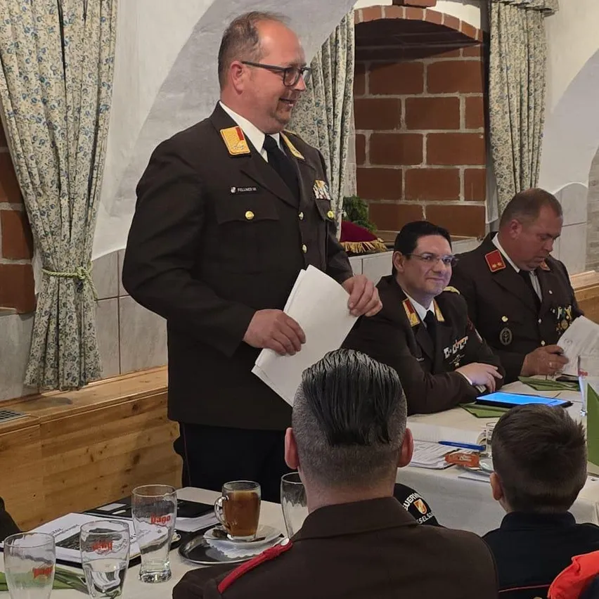 Ein Militäroffizier hält ein Papier in der Hand und spricht eine Gruppe uniformierter Männer an, die an einem Tisch sitzen. Der Raum hat eine Ziegelwand, eine Holzbank und Vorhänge mit Blumenmuster.