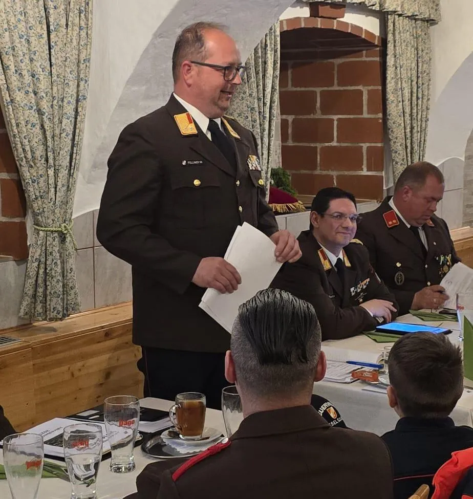 Ein Militäroffizier hält ein Papier in der Hand und spricht eine Gruppe uniformierter Männer an, die an einem Tisch sitzen. Der Raum hat eine Ziegelwand, eine Holzbank und Vorhänge mit Blumenmuster.