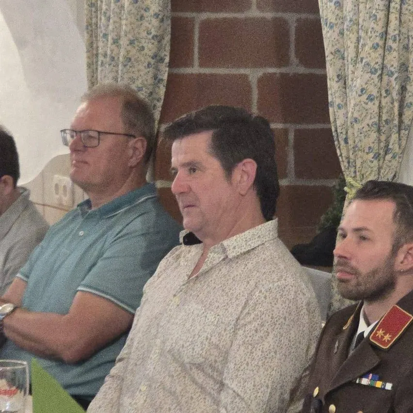 Drei Männer sitzen, einer trägt ein blaues Hemd, ein anderer ein Blumenhemd und der dritte eine Militäruniform. Hinter ihnen befindet sich eine Ziegelmauer mit blumenbedruckten Vorhängen.