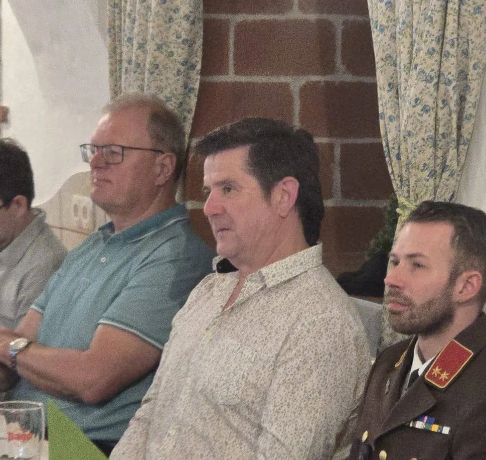 Drei Männer sitzen, einer trägt ein blaues Hemd, ein anderer ein Blumenhemd und der dritte eine Militäruniform. Hinter ihnen befindet sich eine Ziegelmauer mit blumenbedruckten Vorhängen.