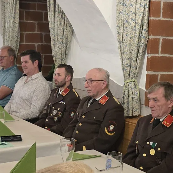 Fünf Männer sitzen an einem Tisch. Drei tragen Militäruniformen, zwei in Zivil. Gläser und Medaillen schmücken die Uniformen. Eine Ziegelmauer und Blumenvorhänge sind im Hintergrund.