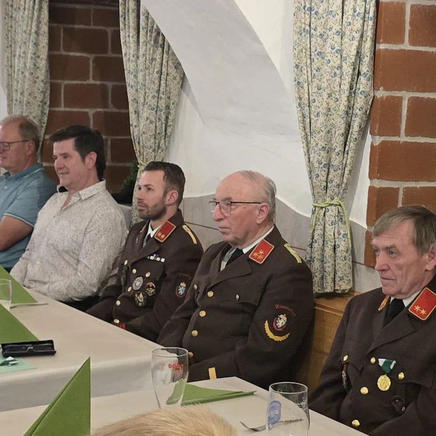 Fünf Männer sitzen an einem Tisch. Drei tragen Militäruniformen, zwei in Zivil. Gläser und Medaillen schmücken die Uniformen. Eine Ziegelmauer und Blumenvorhänge sind im Hintergrund.