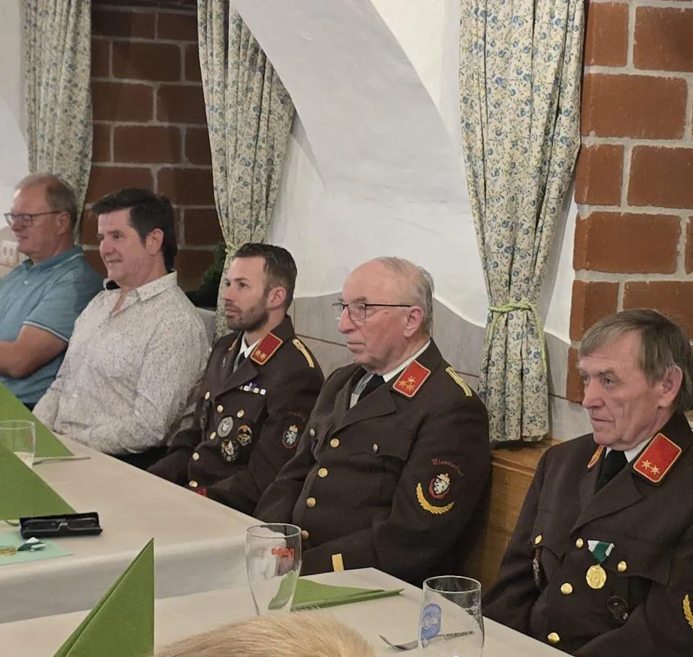 Fünf Männer sitzen an einem Tisch. Drei tragen Militäruniformen, zwei in Zivil. Gläser und Medaillen schmücken die Uniformen. Eine Ziegelmauer und Blumenvorhänge sind im Hintergrund.
