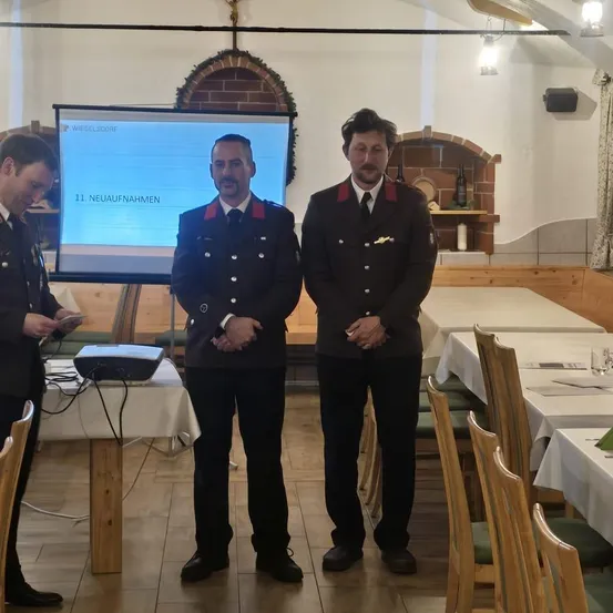 Drei Männer in Uniformen stehen vor einer Leinwand mit der Aufschrift '11 Neueaufnahmen'. Hinter ihnen befindet sich ein Beamer und ein Tisch mit Papieren. Im Hintergrund sind Tische mit Stühlen und ein Kamin zu sehen.