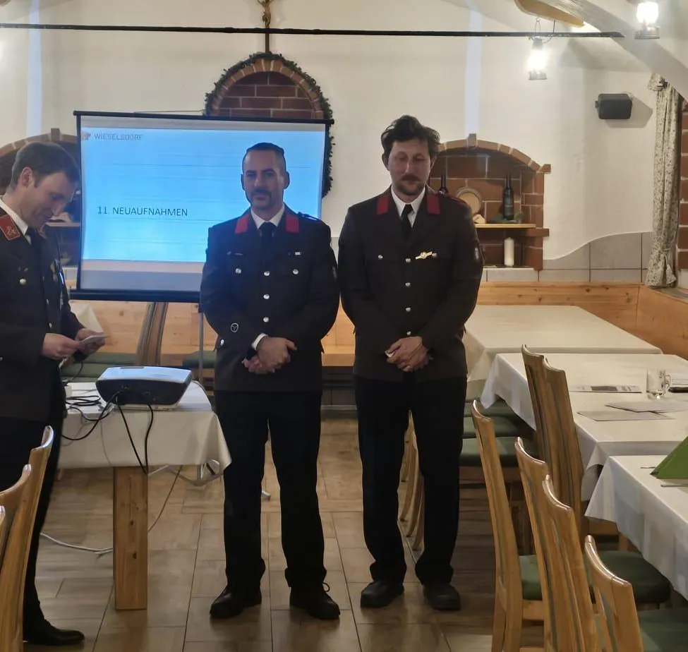 Drei Männer in Uniformen stehen vor einer Leinwand mit der Aufschrift '11 Neueaufnahmen'. Hinter ihnen befindet sich ein Beamer und ein Tisch mit Papieren. Im Hintergrund sind Tische mit Stühlen und ein Kamin zu sehen.