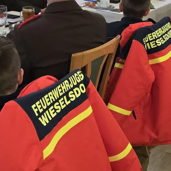 Eine Gruppe Feuerwehrleute in roten Uniformen mit gelben Streifen sitzen um einen Tisch. Die Worte 'Feuerwehrjuges' und 'Wieselsdorf' sind auf ihren Jacken zu sehen.
