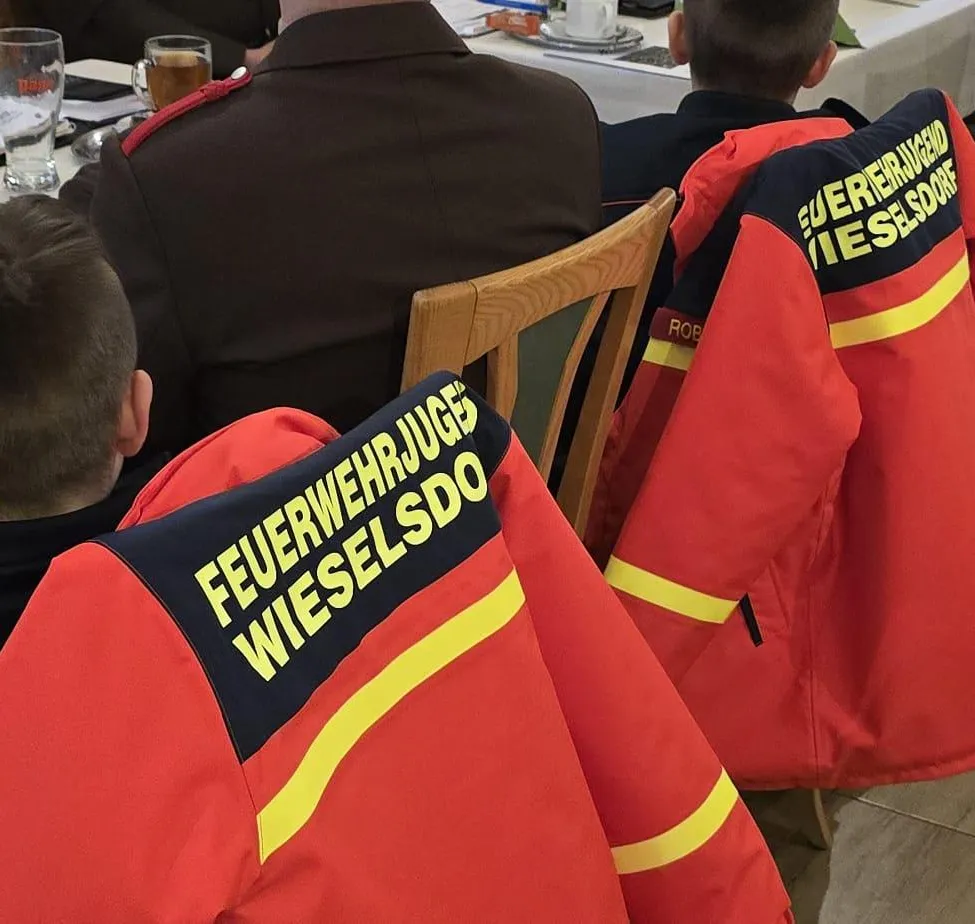 Eine Gruppe Feuerwehrleute in roten Uniformen mit gelben Streifen sitzen um einen Tisch. Die Worte 'Feuerwehrjuges' und 'Wieselsdorf' sind auf ihren Jacken zu sehen.