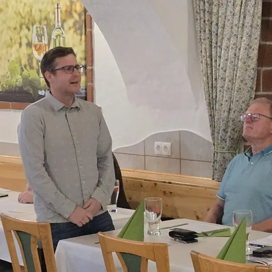 Zwei Männer in einem Restaurant, einer steht und spricht, der andere sitzt. Ein Weinglas und Servietten sind auf dem Tisch. Ein Gemälde mit Flaschen und einem Weinglas hängt an der Wand.
