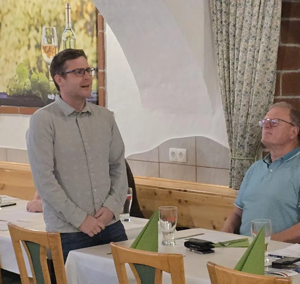 Zwei Männer in einem Restaurant, einer steht und spricht, der andere sitzt. Ein Weinglas und Servietten sind auf dem Tisch. Ein Gemälde mit Flaschen und einem Weinglas hängt an der Wand.