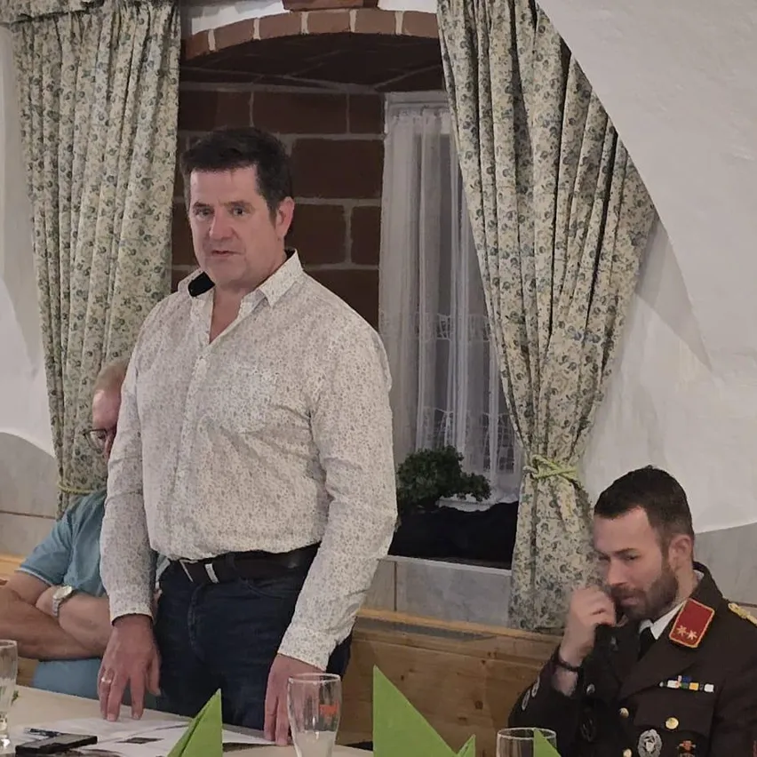 Ein Mann in einem weißen Hemd steht vor einem Tisch mit einem Glas Wasser, während ein weiterer Mann in Uniform hinter ihm sitzt.