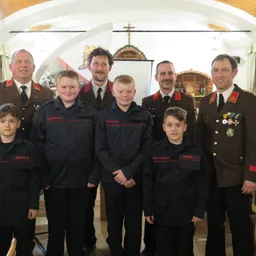 Eine Gruppe von Feuerwehrleuten in Uniform steht in einem Raum, vor ihnen stehen mehrere Kinder. Sie alle lächeln und posieren für ein Foto.
