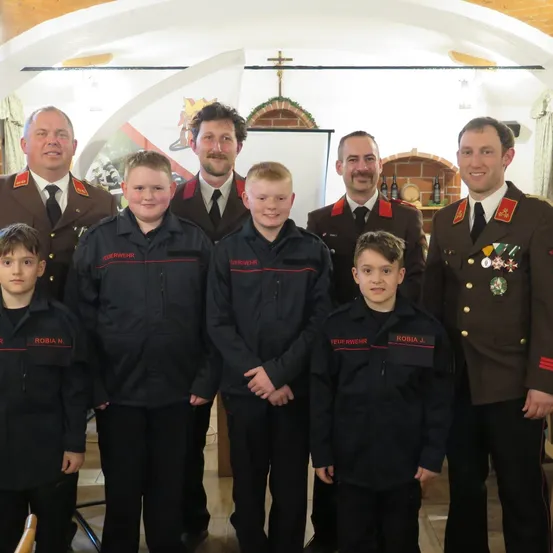 Eine Gruppe von Feuerwehrleuten in Uniform steht in einem Raum, vor ihnen stehen mehrere Kinder. Sie alle lächeln und posieren für ein Foto.