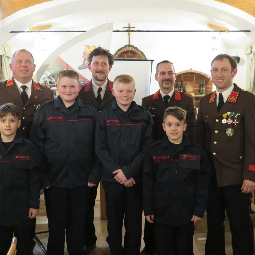 Eine Gruppe von Feuerwehrleuten in Uniform steht in einem Raum, vor ihnen stehen mehrere Kinder. Sie alle lächeln und posieren für ein Foto.