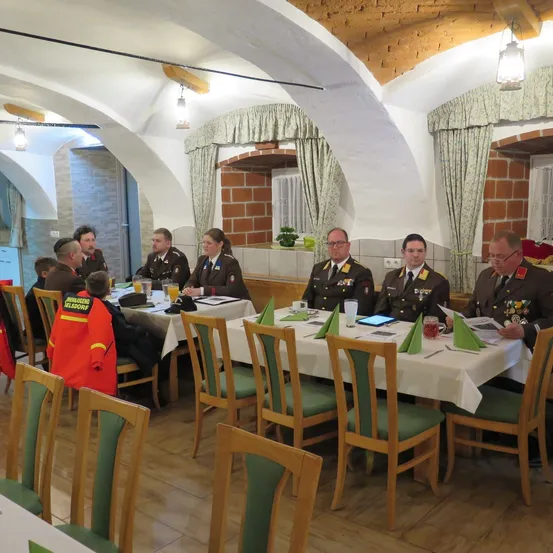 Eine Gruppe uniformierter Militäroffiziere sitzt um einen Tisch in einem Raum mit gewölbten Decken. Ein Kind in einer orangefarbenen Jacke steht neben einem sitzenden Erwachsenen.