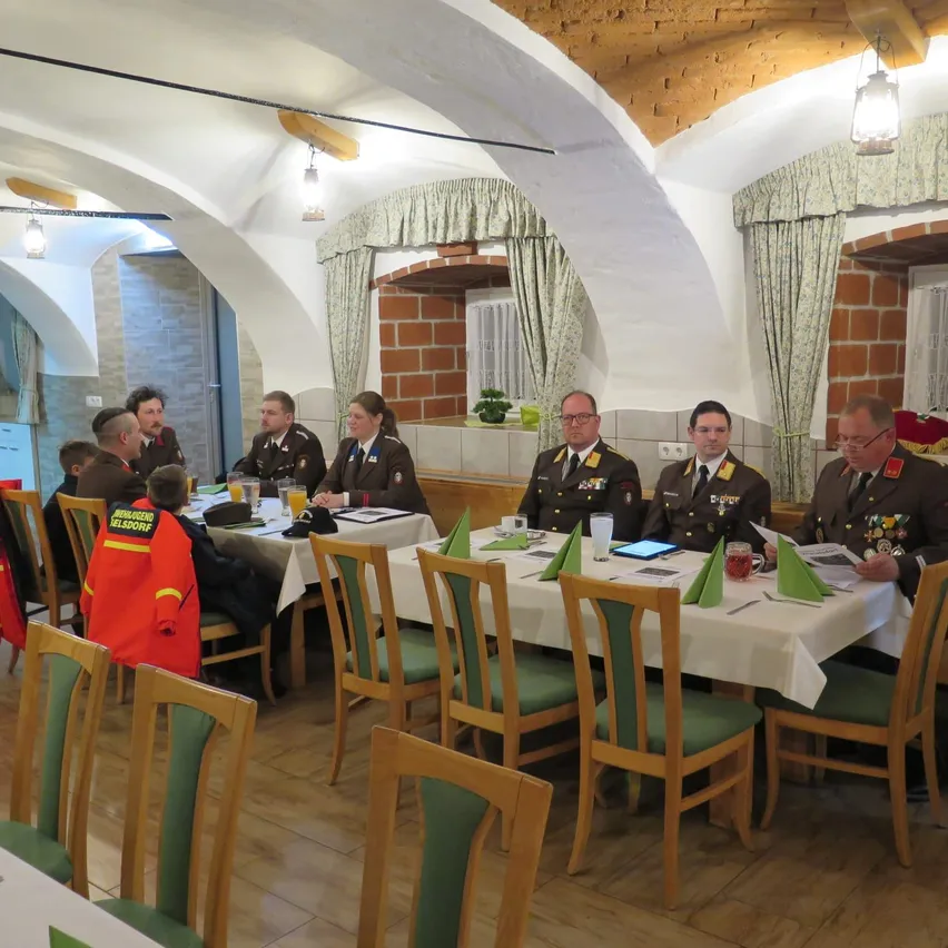 Eine Gruppe uniformierter Militäroffiziere sitzt um einen Tisch in einem Raum mit gewölbten Decken. Ein Kind in einer orangefarbenen Jacke steht neben einem sitzenden Erwachsenen.
