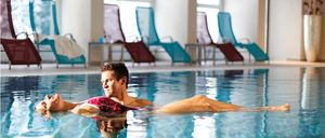 Ein Mann und eine Frau schwimmen in einem Pool und tragen Badeanzüge. Im Hintergrund sind mehrere Liegestühle angeordnet.