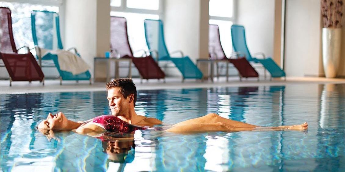 Ein Mann und eine Frau schwimmen in einem Pool und tragen Badeanzüge. Im Hintergrund sind mehrere Liegestühle angeordnet.