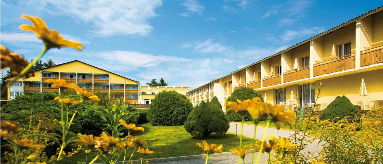 Ein gelbes Gebäude mit Balkonen und Fenstern, umgeben von Büschen und Bäumen unter einem blauen Himmel.