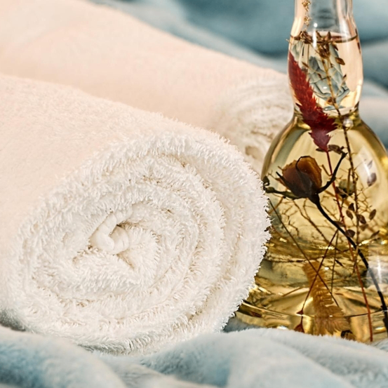 Ein gerolltes weißes Handtuch liegt auf einem blauen Tuch neben einer Flasche ätherischem Öl mit Kräutern darin.