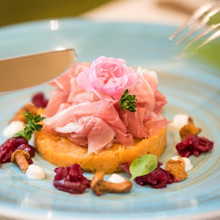 Ein Teller mit einer Scheibe Schinken, einer rosa Blume und einem Pilz. Es hat rote Rüben und Sahne an den Seiten. Ein Messer schneidet durch das Gericht.