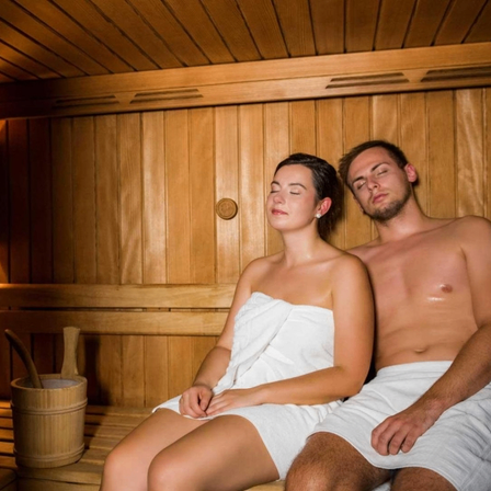 Ein Mann und eine Frau entspannen sich in einer Holzsauna, beide mit Handtüchern um die Hüften. Die Frau sitzt mit geschlossenen Augen, während der Mann neben ihr ruht. Ein Holzeimer und ein Schöpflöffel sind in der Nähe.