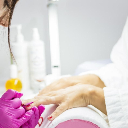 Eine Frau erhält in einem Salon eine Maniküre und trägt pinke Handschuhe. Im Hintergrund befinden sich ein gelbes Objekt und Flaschen.