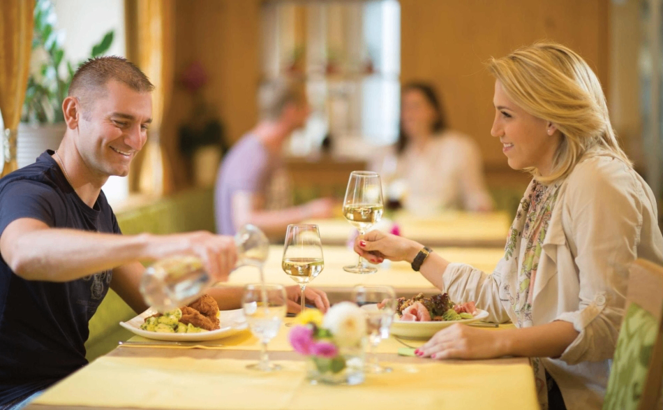 Ein Paar isst in einem Restaurant, lächelt und hält Weingläser in der Hand. Der Tisch ist mit Tellern, Gläsern und einer Blumendekoration gedeckt. Hinter ihnen sitzen ein weiteres Paar und eine Frau, mit einem Fenster im Hintergrund.