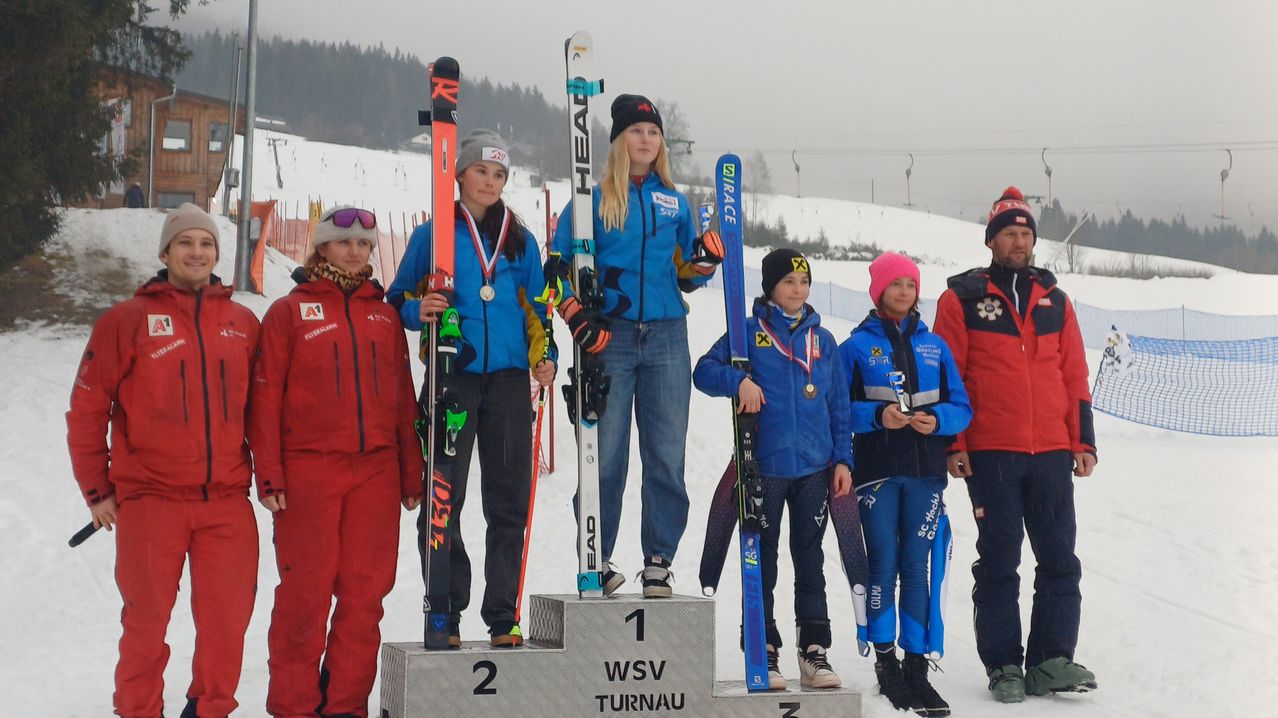 Fünf Skifahrer stehen auf einem Podium mit verschneiten Bergen im Hintergrund. Sie tragen Wintersportausrüstung und halten Skier. Der Podest hat die Zahlen 1, 2 und 3.