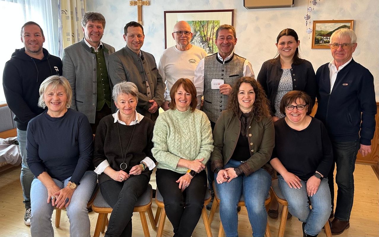 Eine Gruppe von Menschen, sowohl Männer als auch Frauen, lächelt und posiert für ein Foto in einem Raum. Sie tragen alle Freizeitkleidung. Hinter ihnen ist eine Wand mit einem Kreuz und einem Bilderrahmen.