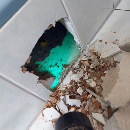 Ein beschädigter Bereich eines Badezimmerbodens zeigt zerbrochene Fliesen und ein Loch in der Wand, durch das eine grüne Substanz austritt.