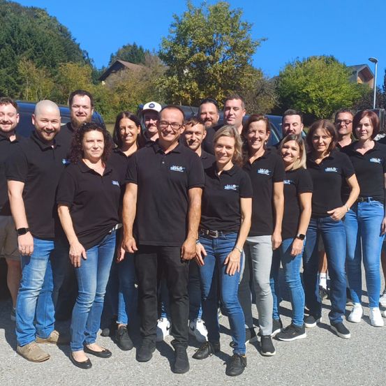 Eine Gruppe von Menschen in schwarzen Polo-Shirts und Jeans posiert für ein Foto vor einem Van, mit Bäumen und Häusern im Hintergrund.