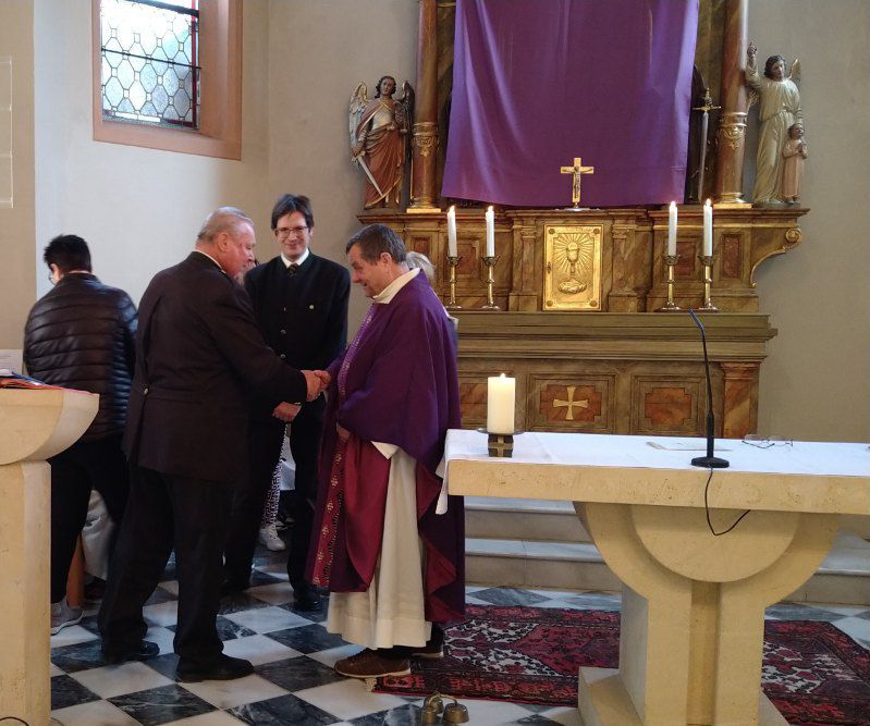 Ein Priester in einer violetten Robe begrüßt einen Mann in einem schwarzen Anzug in einer Kirche. Dahinter steht ein weiterer Mann.