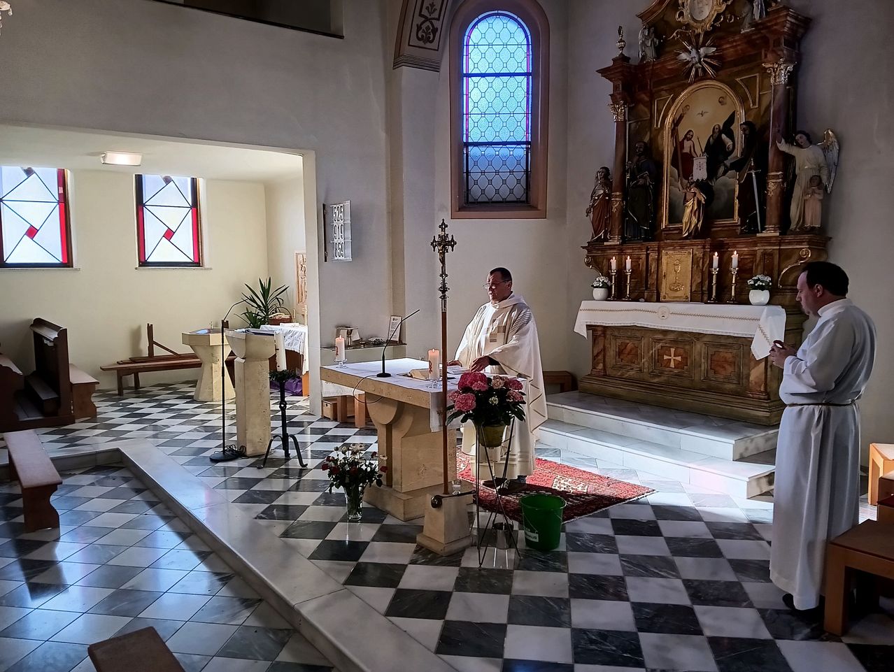 Ein Priester steht vor einem Altar mit Blumen und Kerzen in einer Kirche. Ein Kruzifix befindet sich an der Wand hinter ihm.