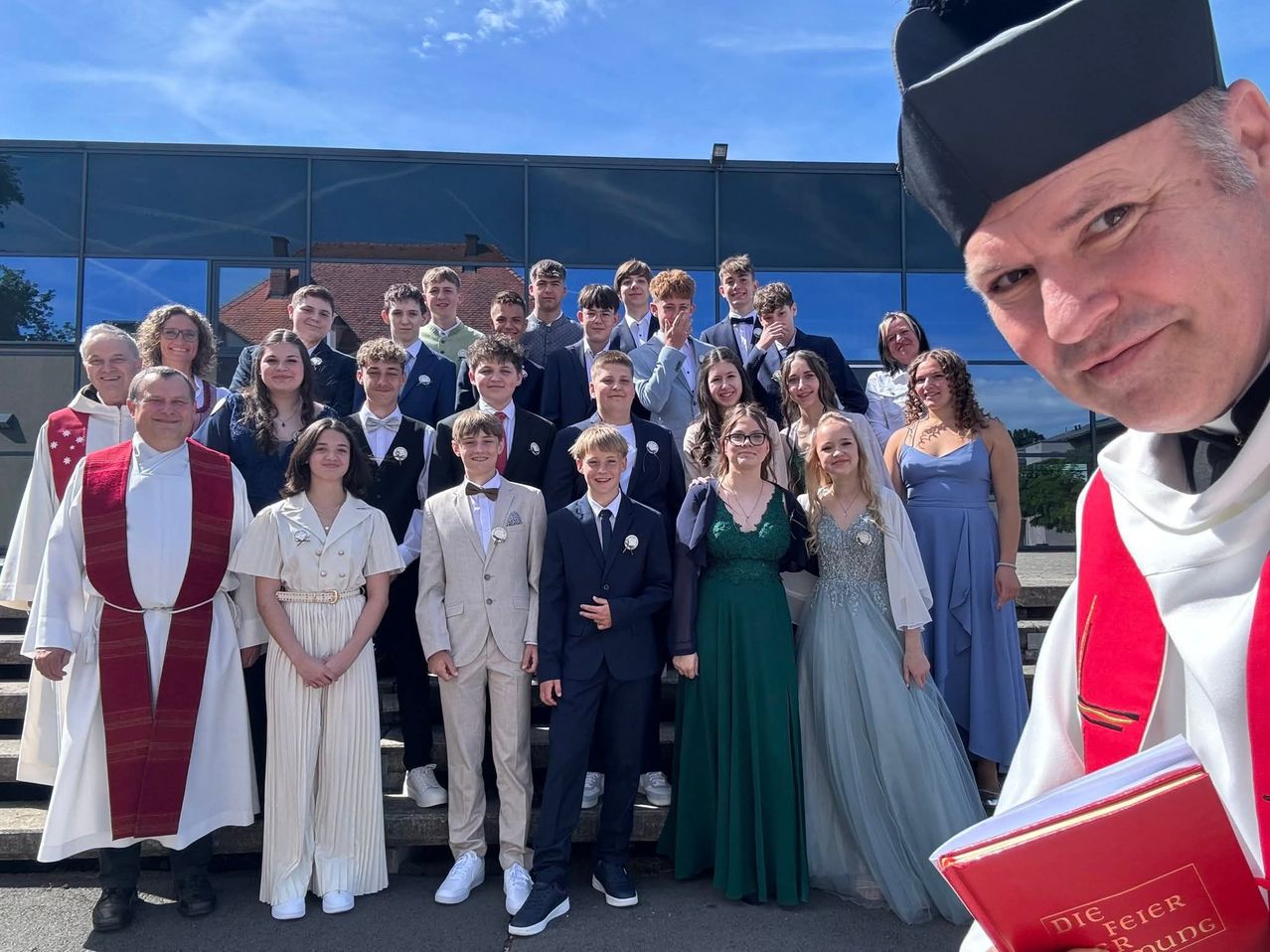 Eine Gruppe formell gekleideter Menschen posiert für ein Foto vor einem Gebäude, wobei ein Priester ein Buch in der Hand hält.
