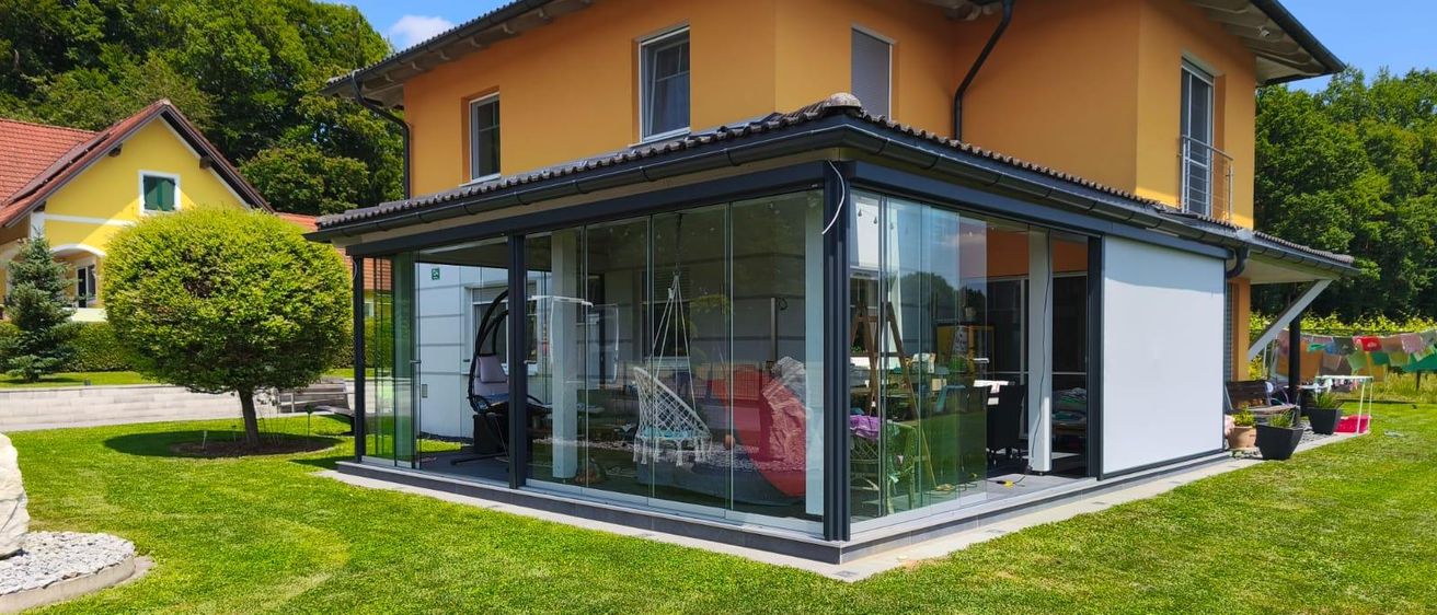 Ein modernes Haus mit einer Glasverlängerung und einem robotergesteuerten Rasenmäher auf dem Rasen. Bäume und ein Baumstumpf sind im Hintergrund sichtbar.