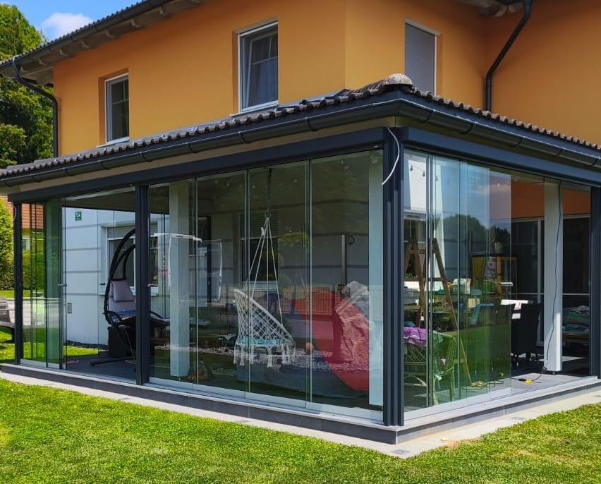 Ein modernes Haus mit einer Glasverlängerung und einem robotergesteuerten Rasenmäher auf dem Rasen. Bäume und ein Baumstumpf sind im Hintergrund sichtbar.