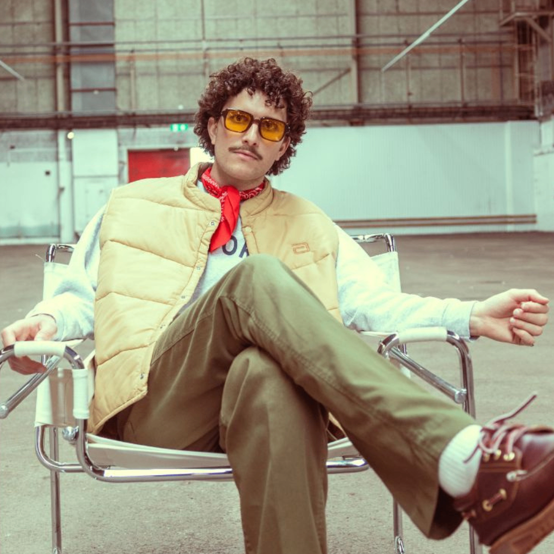 Ein Mann mit lockigem Haar und Brille sitzt in einem Stuhl und trägt eine beige Weste, einen roten Schal und grüne Hosen. Er stützt seine Arme auf den Armlehnen des Stuhls.